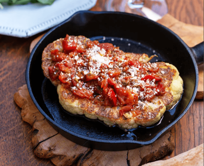 A cauliflower steak resting in a cast iron pan topped with bruschetta and grated cheese